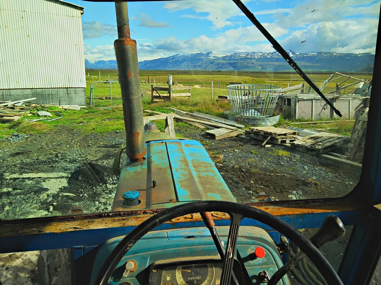 The view from the cab of a rusting tractor across a farmyard, mountains on the horizon