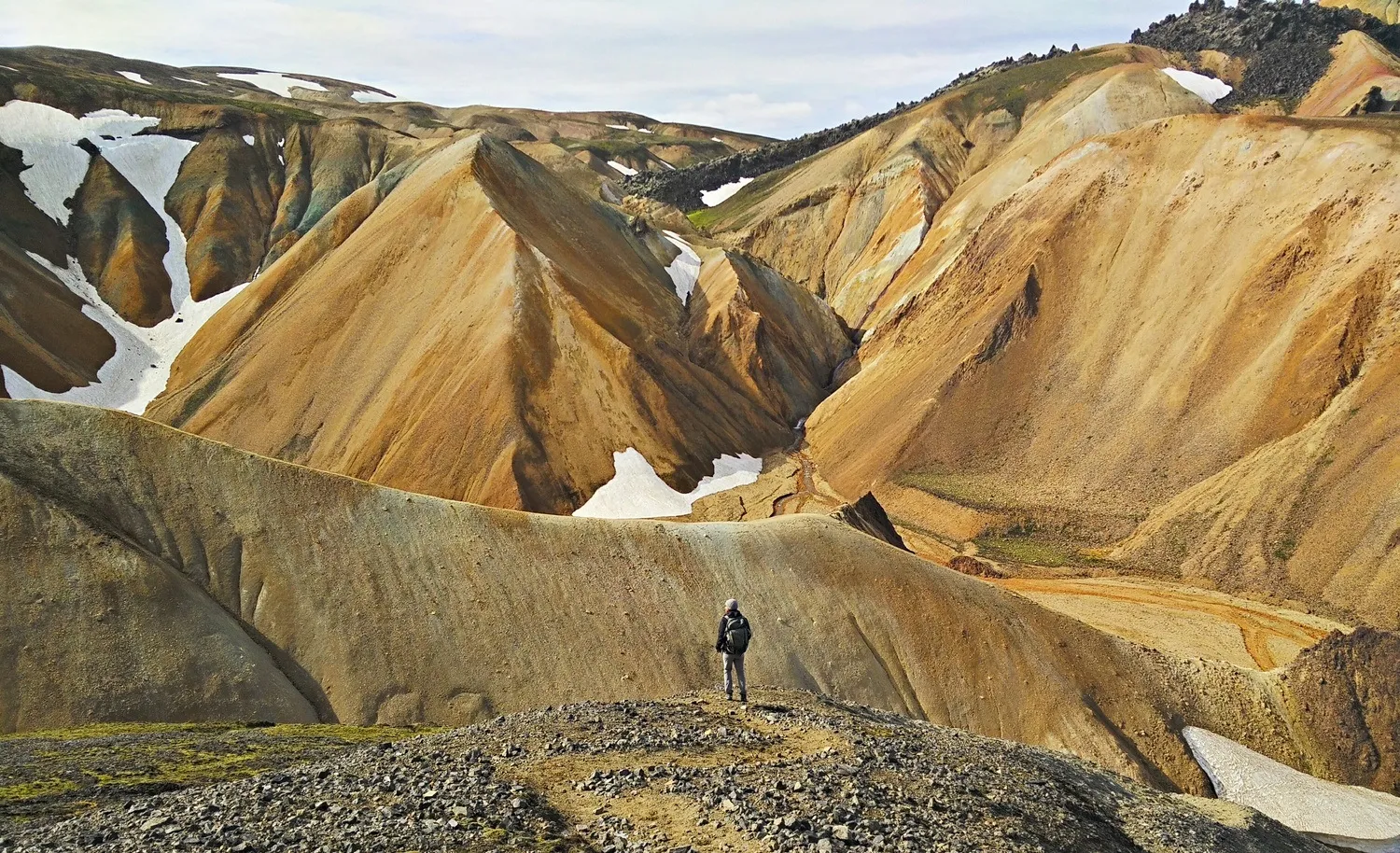 A lone hiker on a ridge among banded rhyolite mountains, snow still lying in the gullies