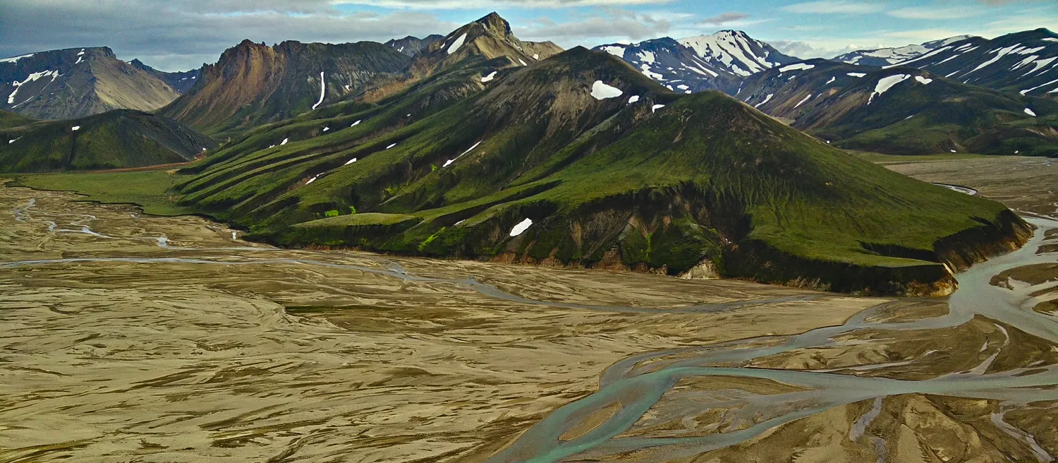 A braided glacial river spreading across a wide valley floor, green mossy slopes and snow-streaked peaks beyond