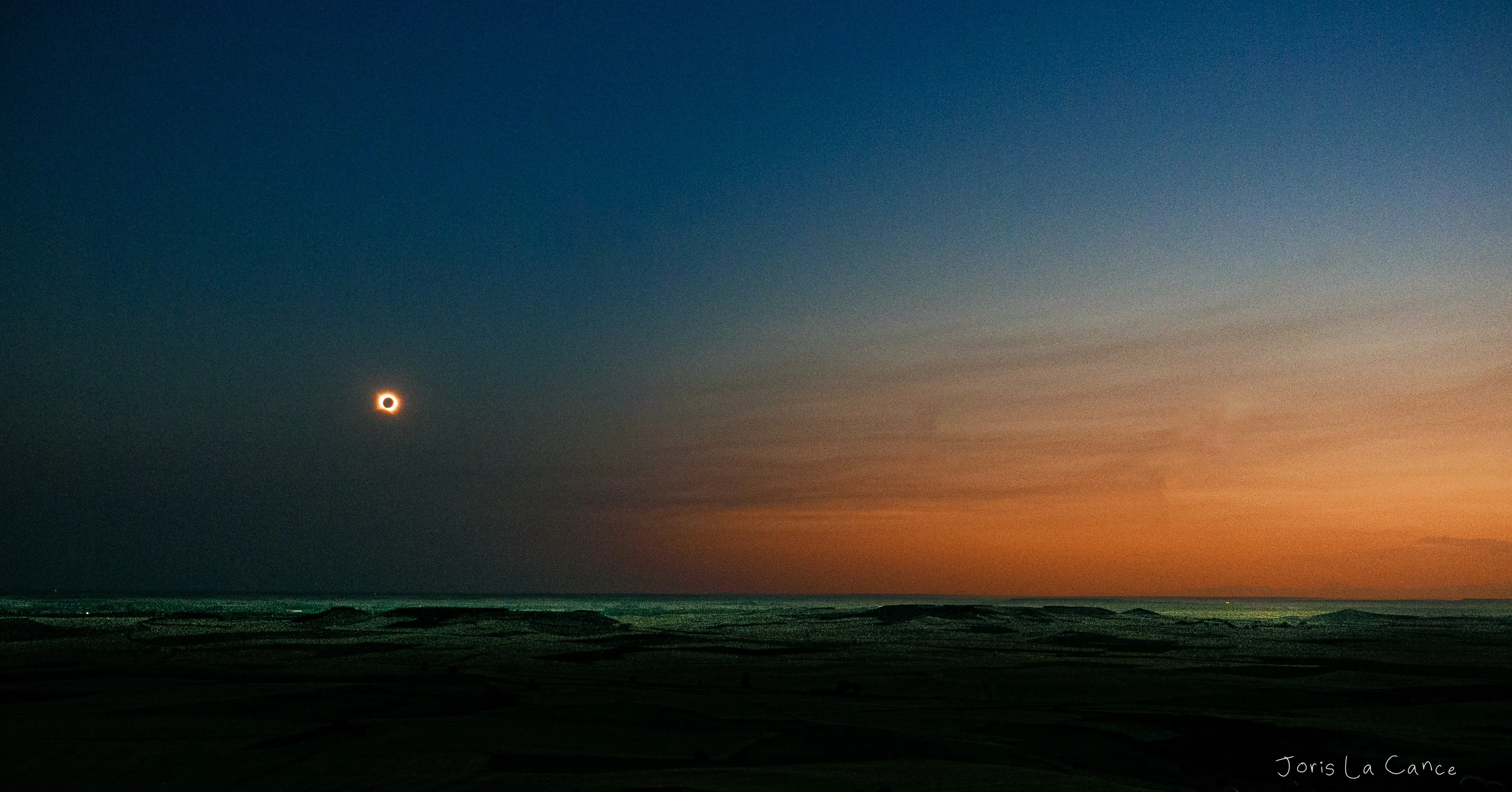 Totality: the eclipsed sun and its corona low over a darkened sea, orange twilight along the horizon