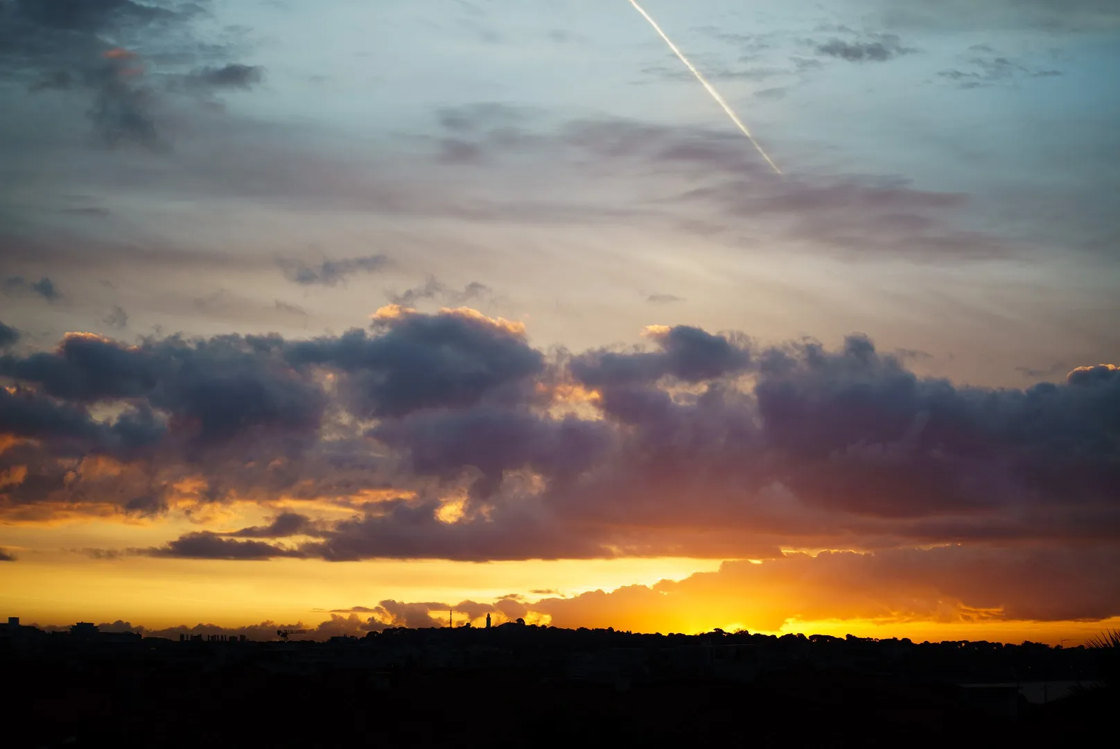 Sunset over a city skyline, layered cloud and a single contrail crossing the glow