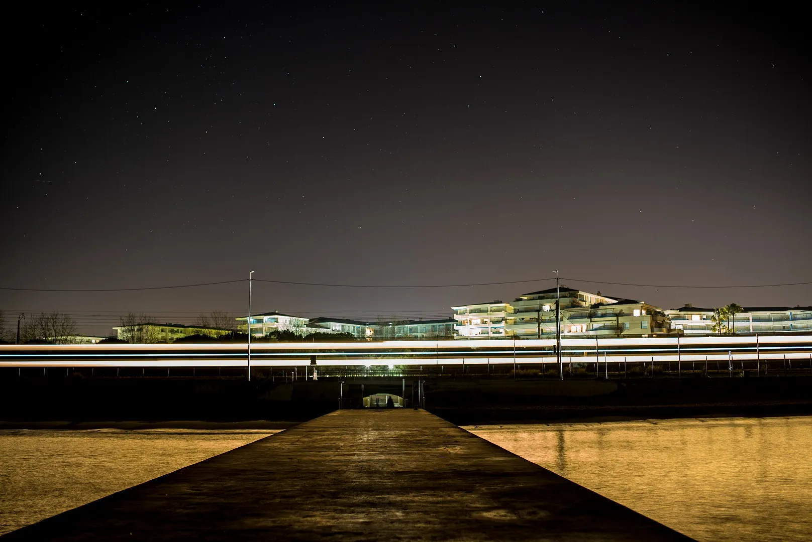 Night long exposure from a jetty across still water to a lit riverside building, traffic drawn as light trails