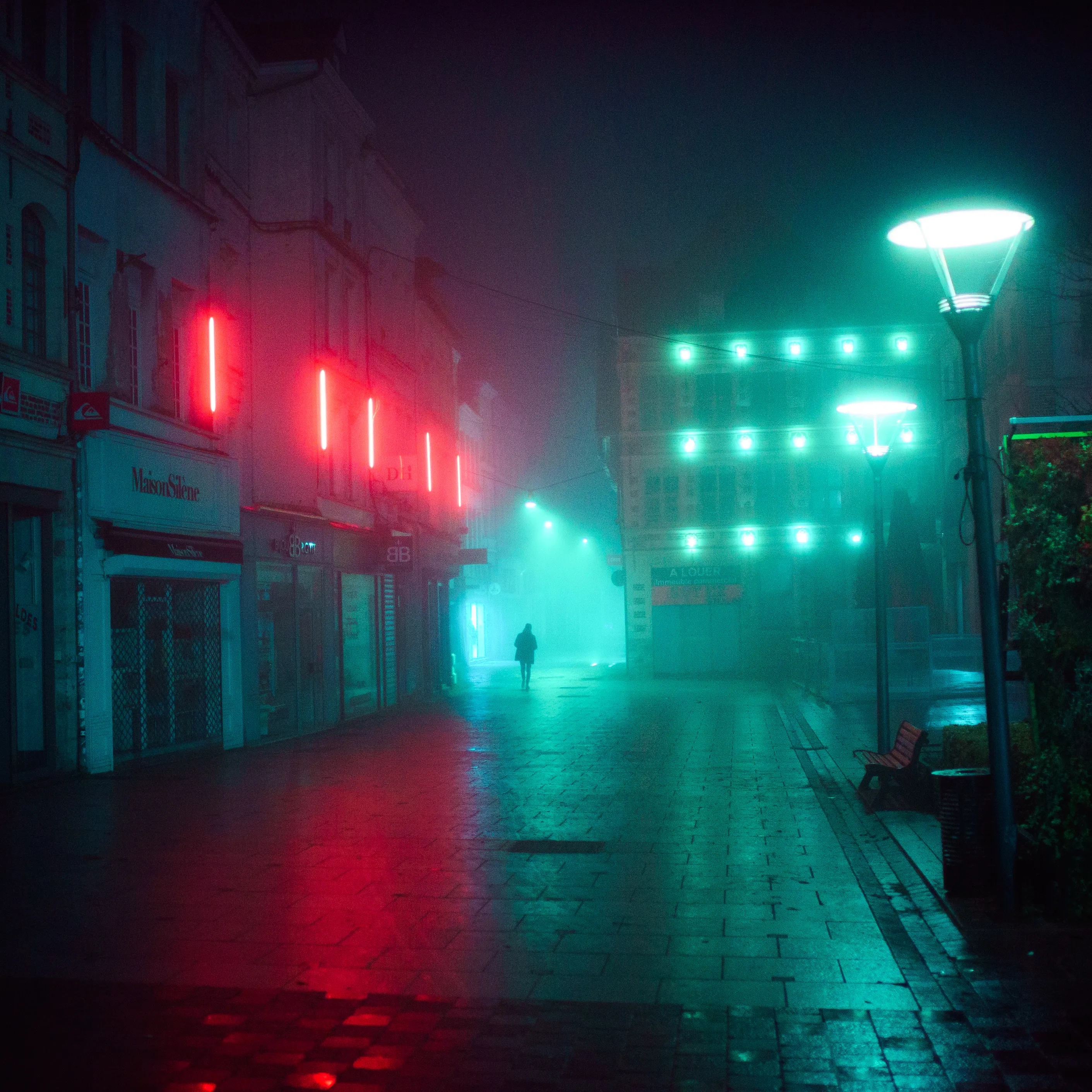 A foggy street at night, red neon on one side and green light ahead, a lone figure on the wet cobbles