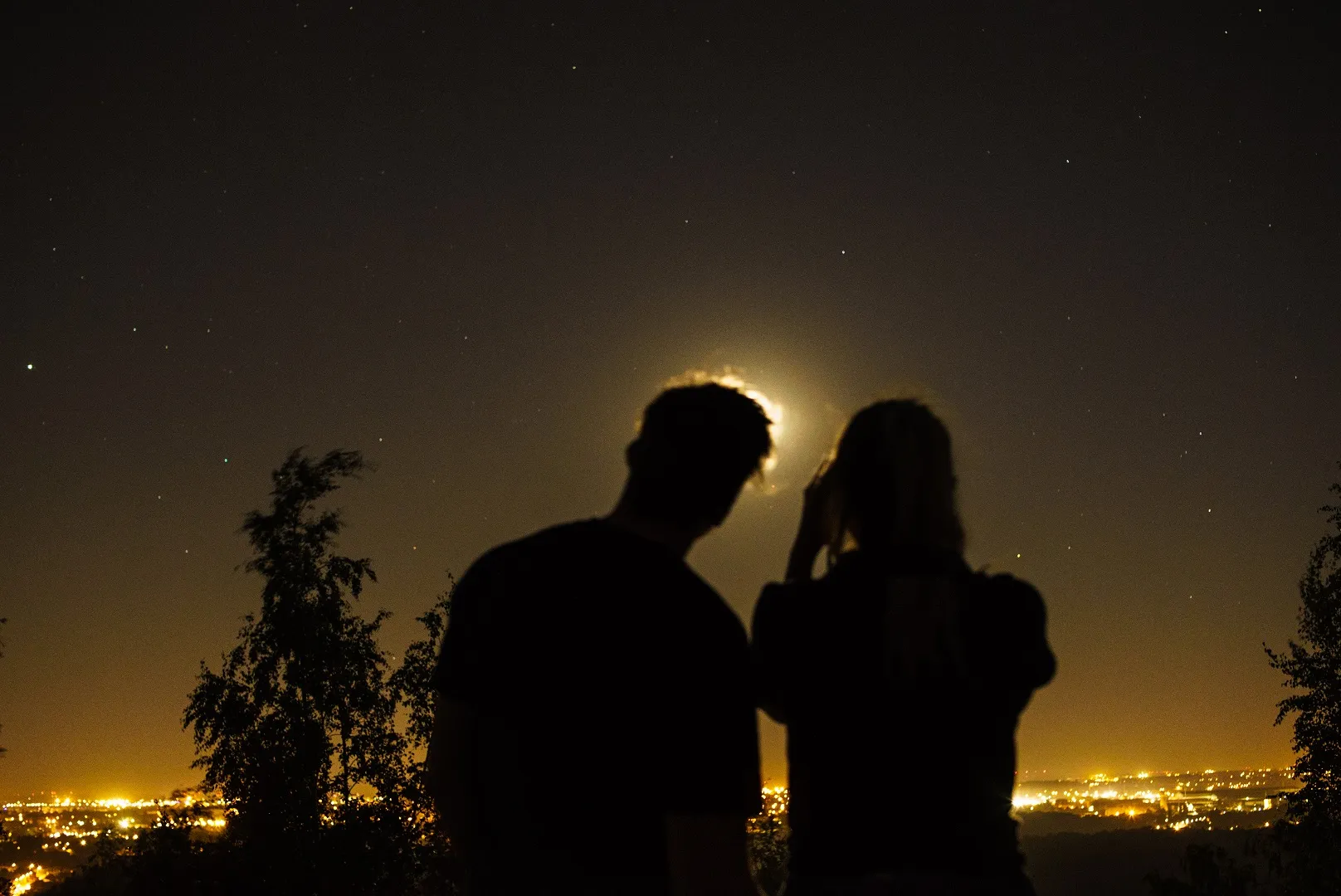 Two people silhouetted on a hillside at night, city lights below and the moon glowing through cloud between them