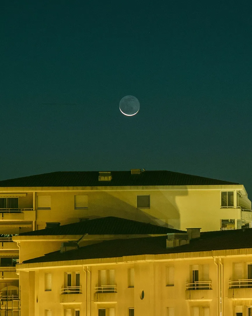 A thin crescent moon in a green-blue twilight sky above apartment rooftops