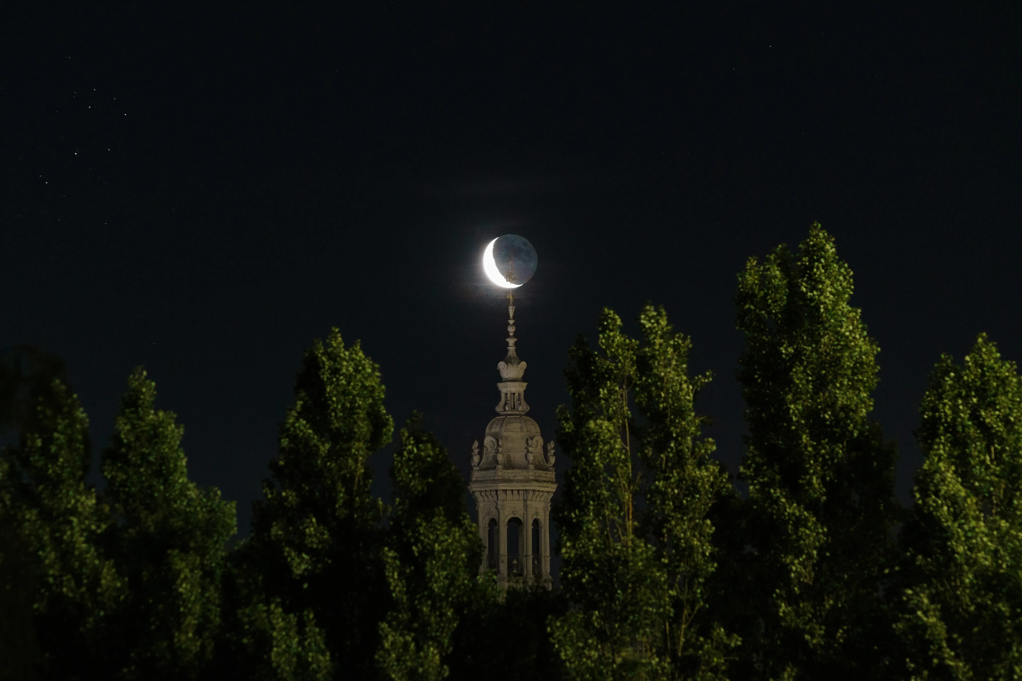 A crescent moon above a church tower framed by poplars at night