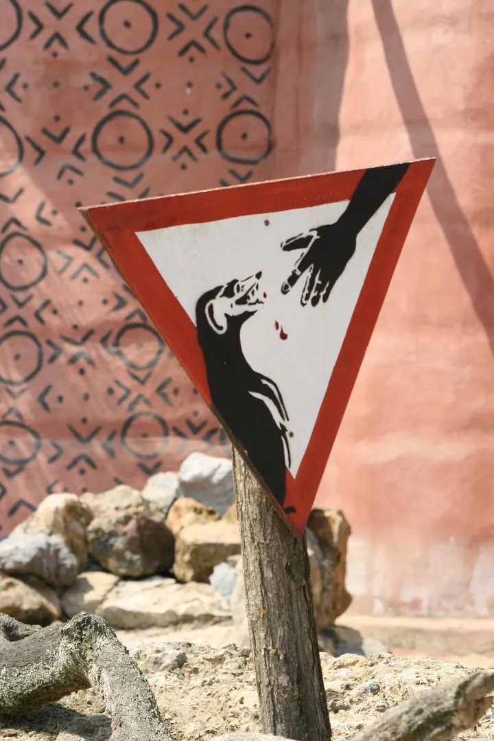 A faded triangular warning sign on a wooden post, showing a hand and falling drops, against a patterned pink wall
