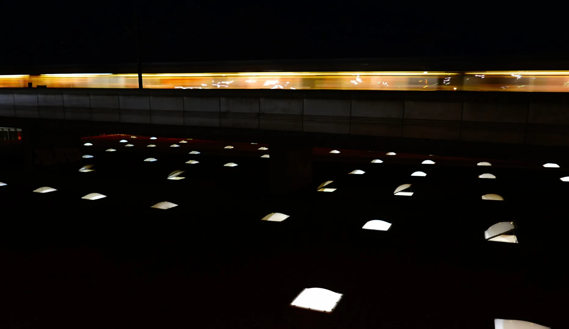 Long exposure at night: a tram streaks above a plaza set with rows of glowing ground lights