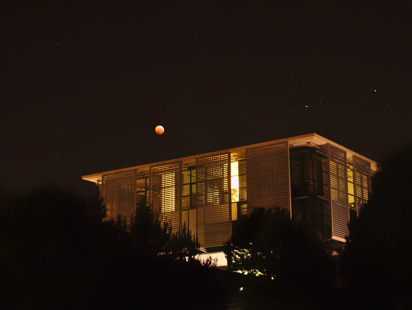 A deep orange moon low in the night sky above a building with a lit louvred facade