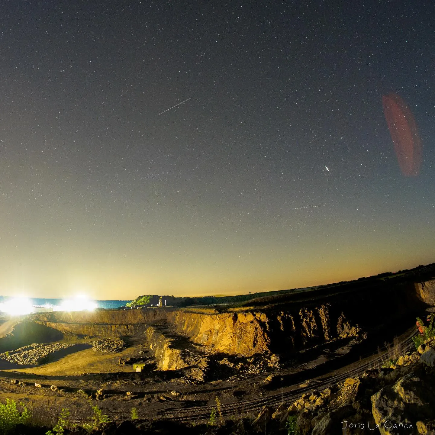 A fisheye view of a floodlit quarry at night with meteors crossing the sky