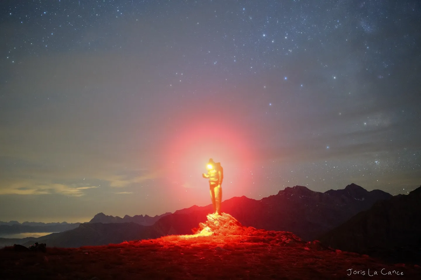 A figure on a summit lit by a red headlamp, the Milky Way overhead